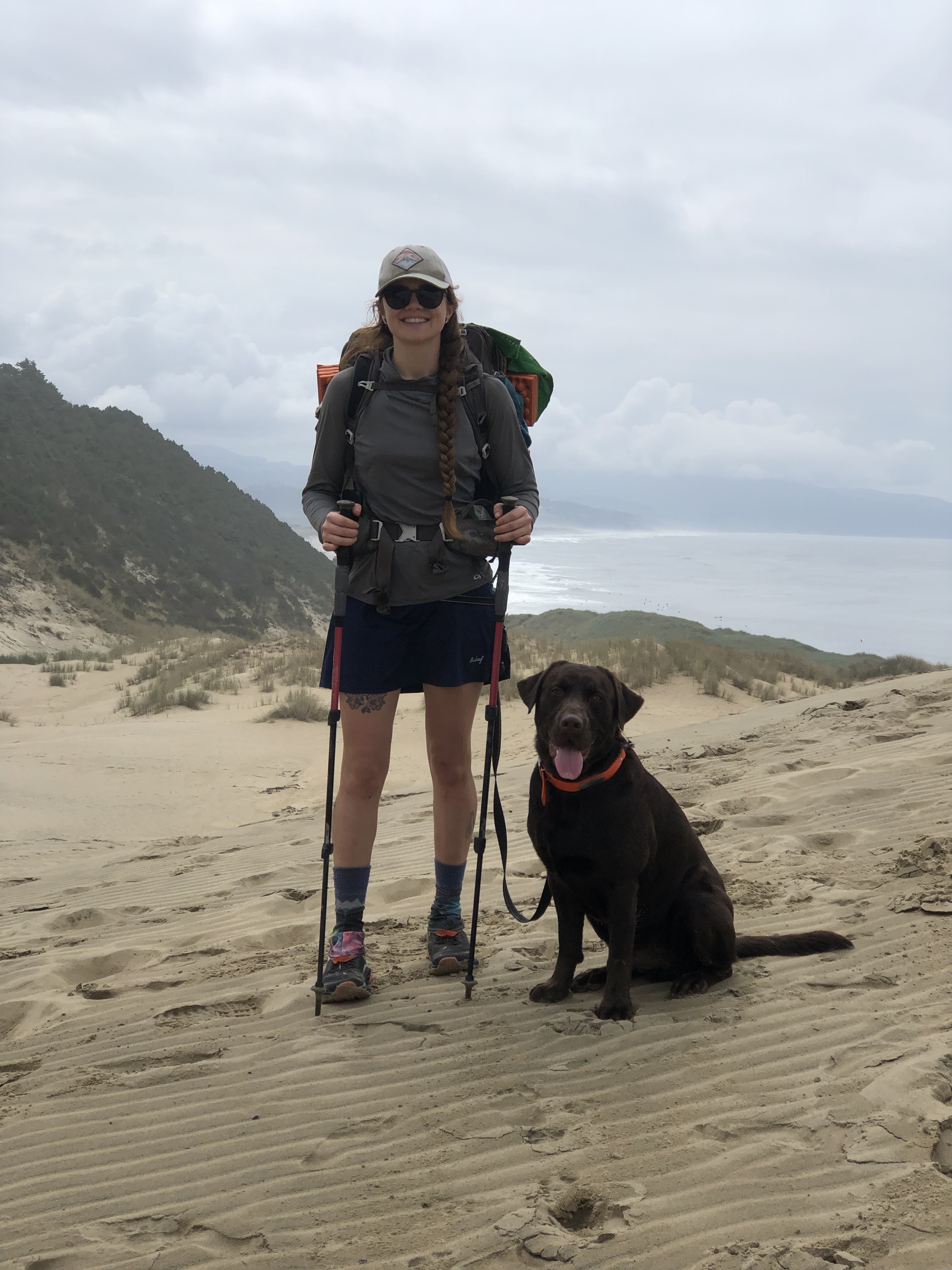 Woman with long braid,backpacking backpack, sunglasses, hat and hiking pole with large brown dog on sand dune overlooking ocean